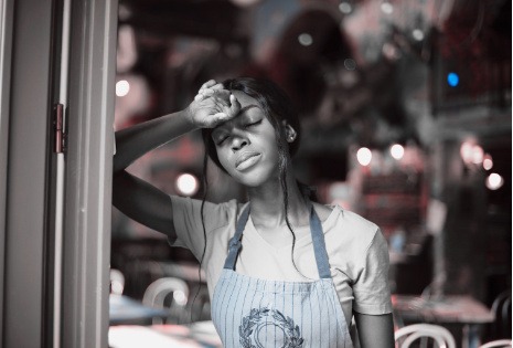 Aidante fatiguée appuyée contre son bras, les yeux fermés près d’une fenêtre, exprimant l’épuisement émotionnel